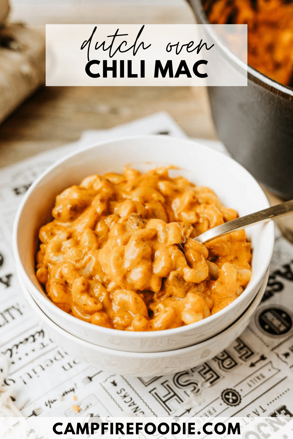 A white bowl filled with creamy chili mac sits on a patterned surface with a spoon inside. Behind it is a black Dutch oven. Text on the image reads dutch oven chili mac and campfirefoodie.com.