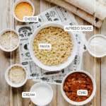 Overhead view of bowls containing macaroni pasta, Alfredo sauce, shredded cheese, cream, and chili, arranged on a wooden table with birch logs. Each bowl is labeled with its contents.