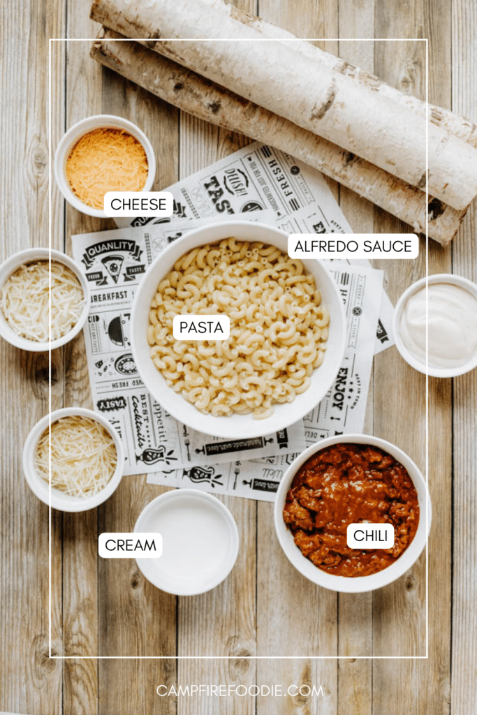 Overhead view of bowls containing macaroni pasta, Alfredo sauce, shredded cheese, cream, and chili, arranged on a wooden table with birch logs. Each bowl is labeled with its contents.