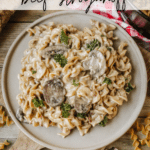 A plate of creamy beef stroganoff with noodles, mushrooms, and herbs sits on a rustic table with uncooked pasta, a red-checkered cloth, and a wooden spoon nearby. Text reads Dutch Oven Beef Stroganoff and campfirefoodie.com.