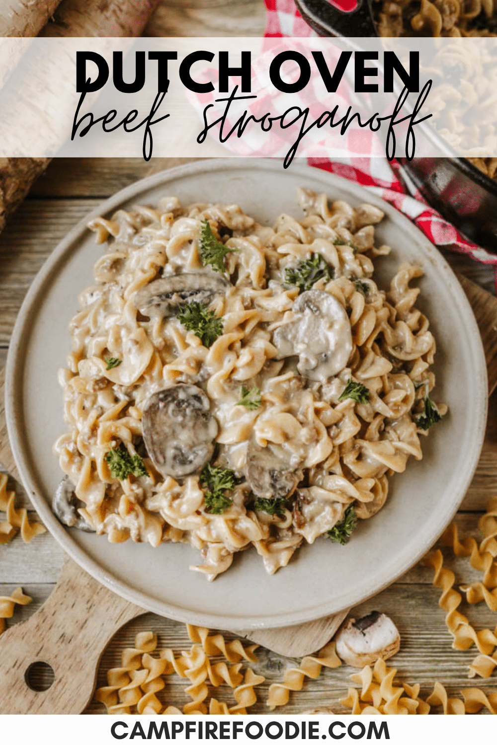 A plate of creamy beef stroganoff with noodles, mushrooms, and herbs sits on a rustic table with uncooked pasta, a red-checkered cloth, and a wooden spoon nearby. Text reads Dutch Oven Beef Stroganoff and campfirefoodie.com.