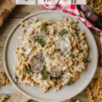 A plate of creamy beef stroganoff with mushrooms and pasta sits on a wooden board next to scattered uncooked noodles. A Dutch oven and a red checked cloth are partially visible in the background.