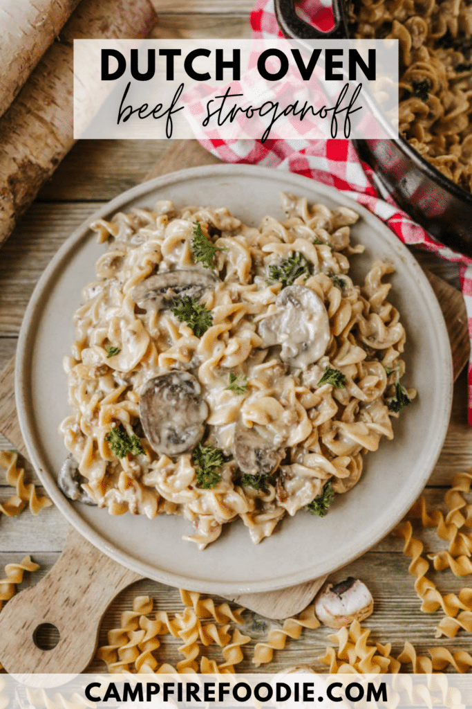 A plate of creamy beef stroganoff with mushrooms and pasta sits on a wooden board next to scattered uncooked noodles. A Dutch oven and a red checked cloth are partially visible in the background.