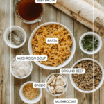 Top-down view of labeled ingredients on a wooden surface: beef broth, pasta, mushroom soup, ground beef, garlic, mushrooms, dried herbs, and chopped green herbs, with the website campfirefoodie.com at the bottom.