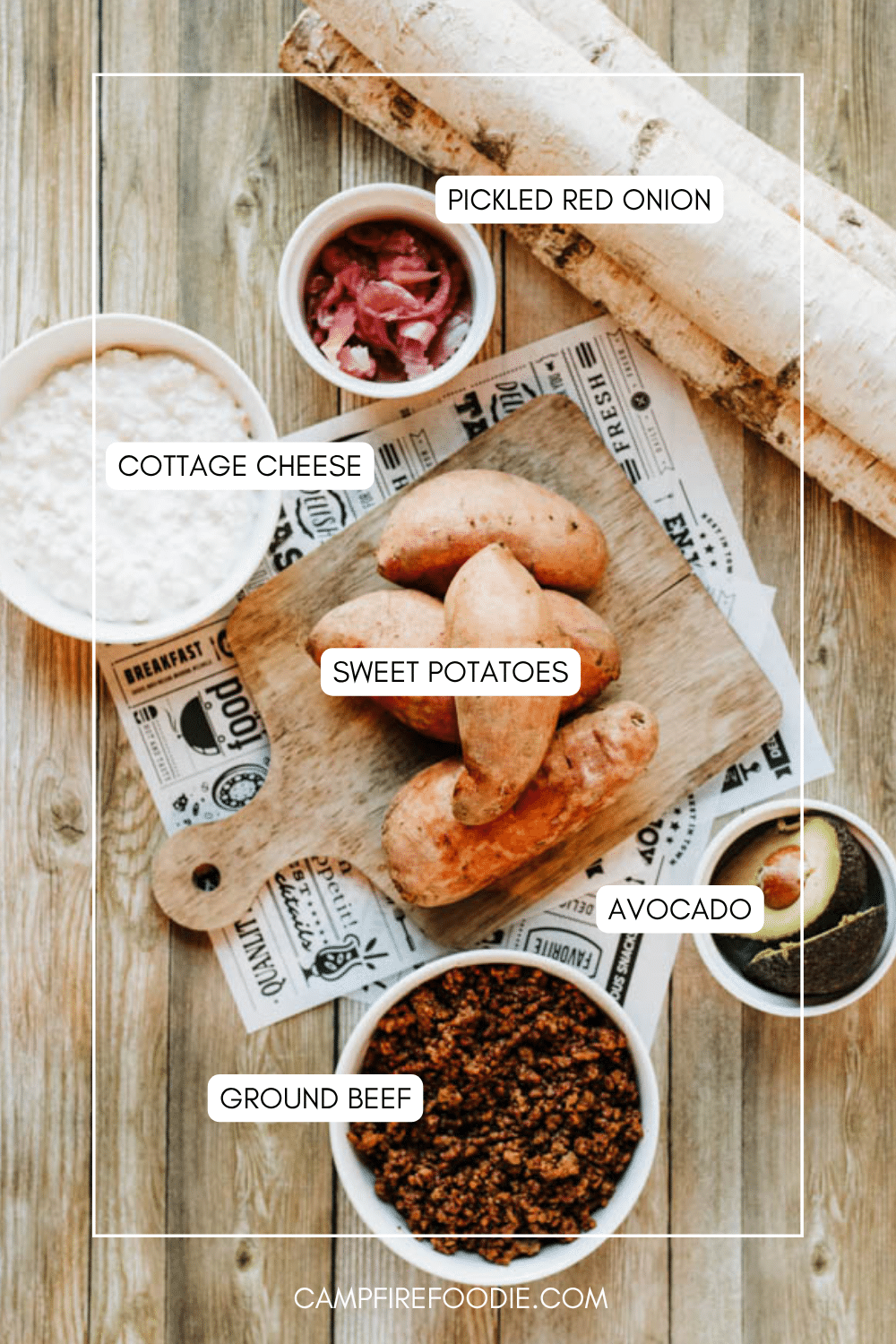 A flat lay of food ingredients on a wooden surface, including sweet potatoes on a cutting board, bowls of ground beef, pickled red onion, cottage cheese, and sliced avocado, with a baguette in the corner.