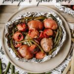 A foil packet filled with cooked sausage, green beans, and potatoes sits on a plate. Text reads Foil Packet Sausage & Green Beans with campfirefoodie.com at the bottom.