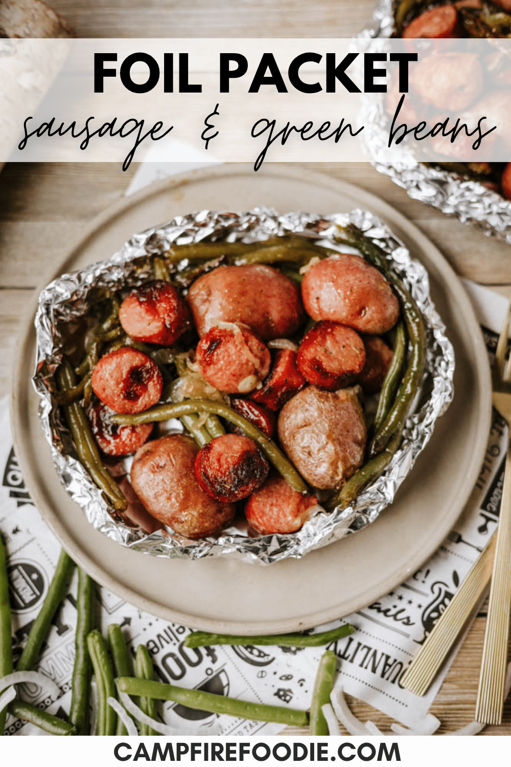 A foil packet filled with cooked sausage, green beans, and potatoes sits on a plate. Text reads Foil Packet Sausage & Green Beans with campfirefoodie.com at the bottom.