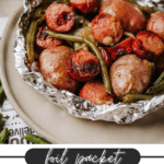 A foil packet filled with cooked sausage slices, green beans, and whole baby potatoes sits on a beige plate. Text below reads, foil packet sausage & green beans campfirefoodie.com.