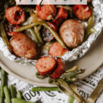 A foil packet filled with sliced sausage, whole baby potatoes, and green beans on a plate. Text on the image reads: Foil Packet Sausage & Beans and campfirefoodie.com.