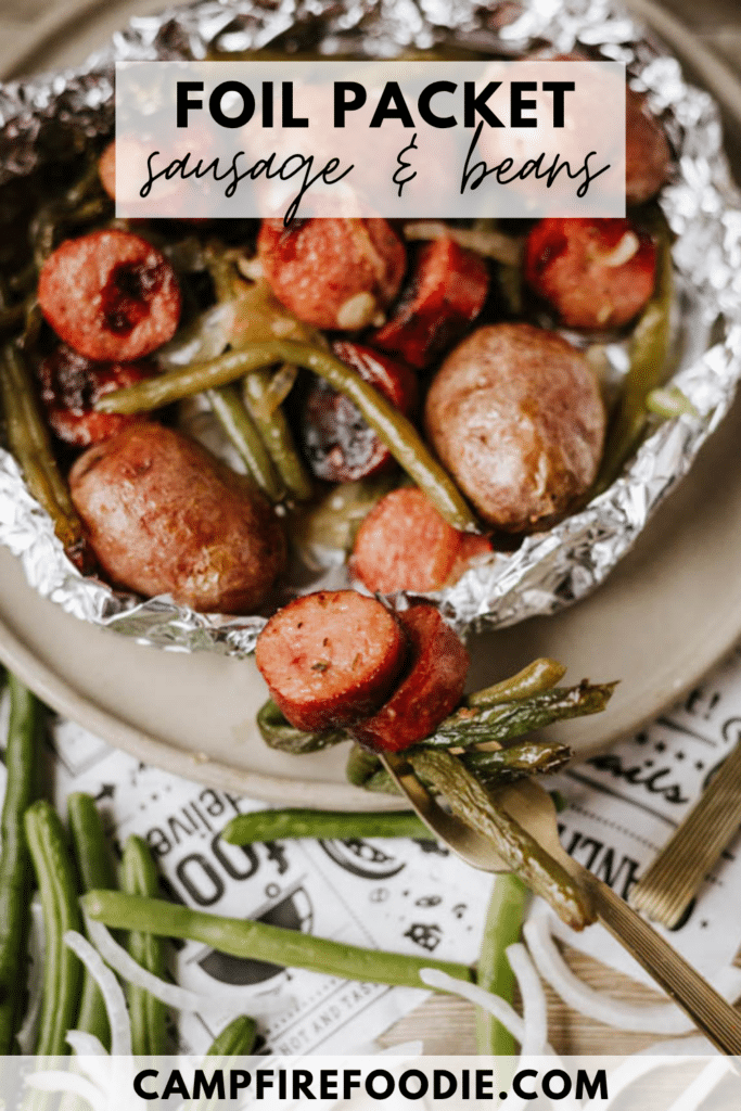 A foil packet filled with sliced sausage, whole baby potatoes, and green beans on a plate. Text on the image reads: Foil Packet Sausage & Beans and campfirefoodie.com.