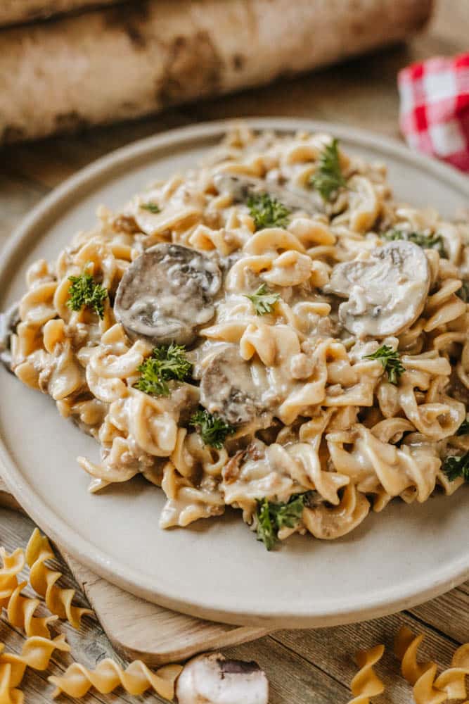 A plate of creamy mushroom pasta, inspired by Dutch oven beef stroganoff, sits on a wooden surface. Spiral noodles are coated in rich sauce with visible mushroom slices. Uncooked pasta and a checkered cloth are nearby.