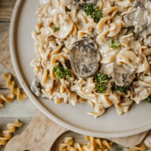 A plate of creamy mushroom pasta with rotini noodles, inspired by Dutch oven beef stroganoff, is garnished with fresh parsley and sits on a wooden board with uncooked pasta spirals scattered around.