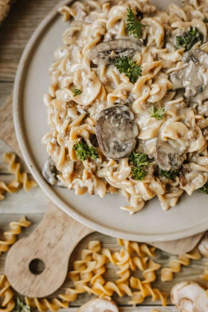 A plate of creamy mushroom pasta with rotini noodles, inspired by Dutch oven beef stroganoff, is garnished with fresh parsley and sits on a wooden board with uncooked pasta spirals scattered around.