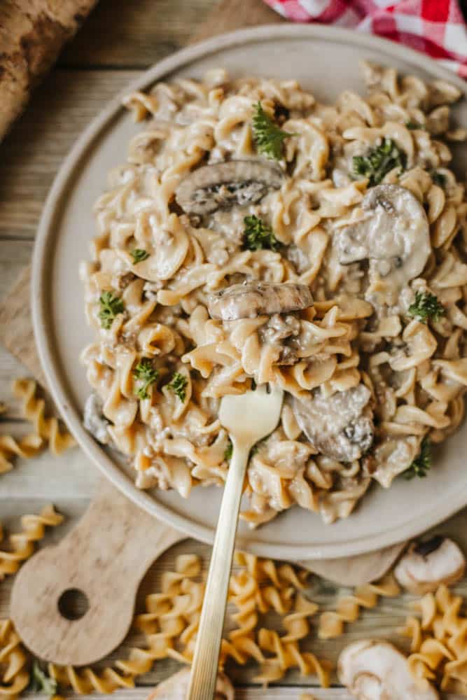 A plate of creamy mushroom pasta, reminiscent of dutch oven beef stroganoff, is garnished with parsley on a wooden board; a gold fork rests on the plate. Uncooked pasta spirals and mushroom slices are scattered nearby.