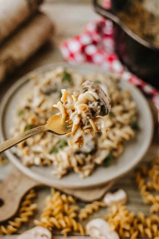 A close-up of a fork holding a bite of creamy pasta with mushrooms over a full plate of pasta, reminiscent of dutch oven beef stroganoff, surrounded by uncooked rotini and fresh mushroom slices on a wooden surface.