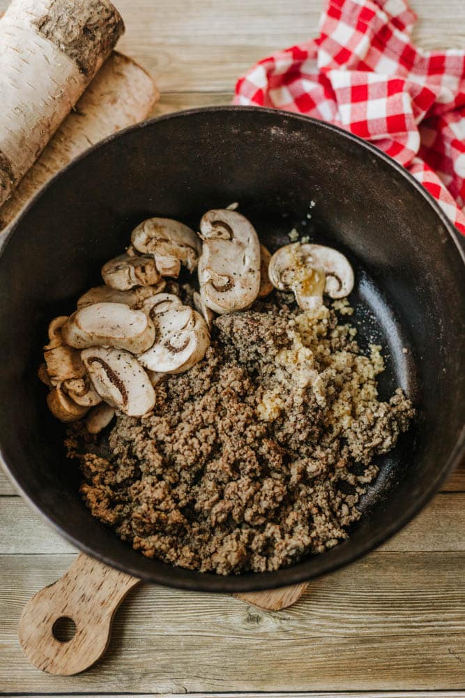 Ground meat, sliced mushrooms, and minced garlic simmer in a black pot, perfect for a hearty dutch oven beef stroganoff. The pot sits on a wooden surface with a red and white checkered cloth in the background.