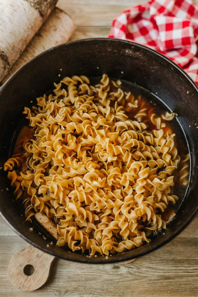 A black pot, perfect for dutch oven beef stroganoff, is filled with uncooked spiral pasta noodles sitting in broth on a wooden surface, with a red and white checkered cloth in the background.