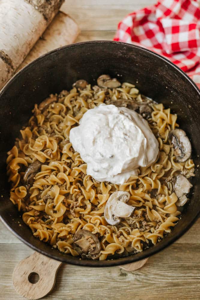 A Dutch oven beef stroganoff featuring cooked egg noodles, sliced mushrooms, and ground meat, topped with a generous dollop of sour cream, sits on a wooden surface next to a red and white checkered cloth.