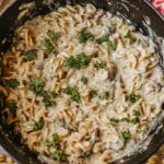 A pot of creamy pasta with mushrooms, inspired by Dutch oven beef stroganoff, sits on a wooden table next to a red and white checkered cloth. Uncooked pasta and mushroom slices are scattered nearby.
