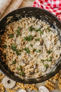 A pot of creamy pasta with mushrooms, inspired by Dutch oven beef stroganoff, sits on a wooden table next to a red and white checkered cloth. Uncooked pasta and mushroom slices are scattered nearby.