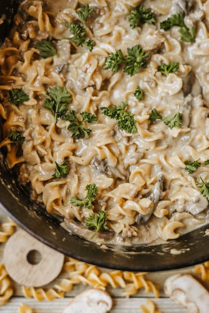 A creamy noodle dish with mushrooms and curly egg noodles, similar to a dutch oven beef stroganoff, garnished with fresh parsley in a black skillet. Uncooked noodles and mushroom slices are partially visible on the table nearby.