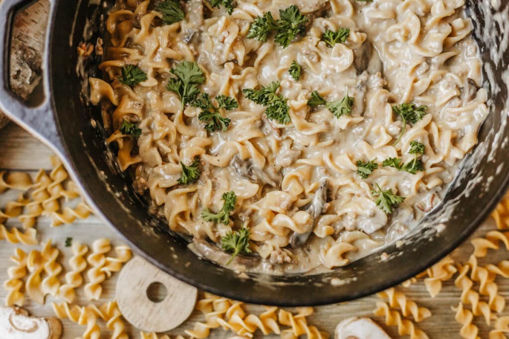 A pot of creamy pasta with rotini noodles, reminiscent of a Dutch oven beef stroganoff, is garnished with fresh parsley and sits on a wooden surface scattered with dry pasta and mushroom slices.