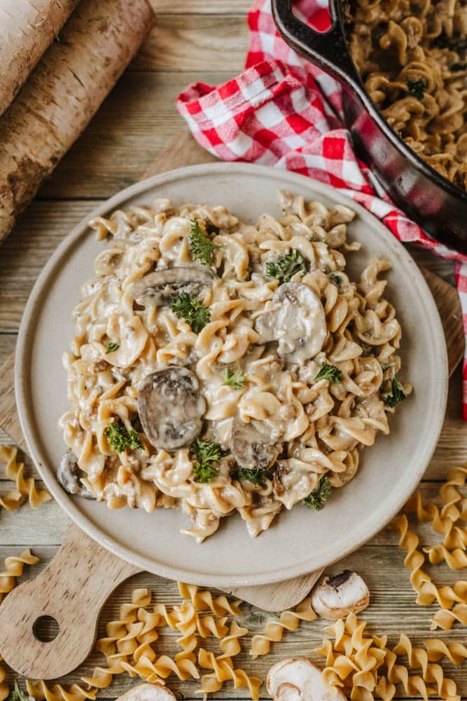 A plate of creamy mushroom pasta garnished with parsley sits on a wooden board, evoking the comfort of dutch oven beef stroganoff, surrounded by uncooked noodles and a red checkered cloth, with a cast iron skillet partially visible in the background.
