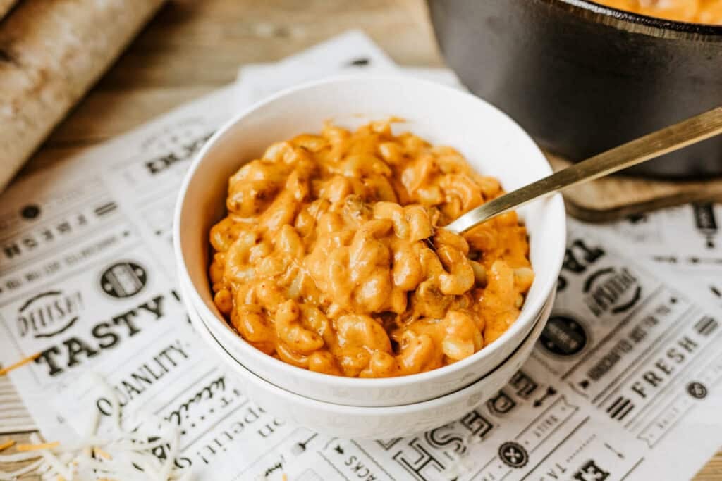 A bowl of creamy macaroni and cheese with a golden-orange sauce sits on a decorative paper, with a spoon in the bowl and a pot of hearty Dutch oven chili mac in the background.