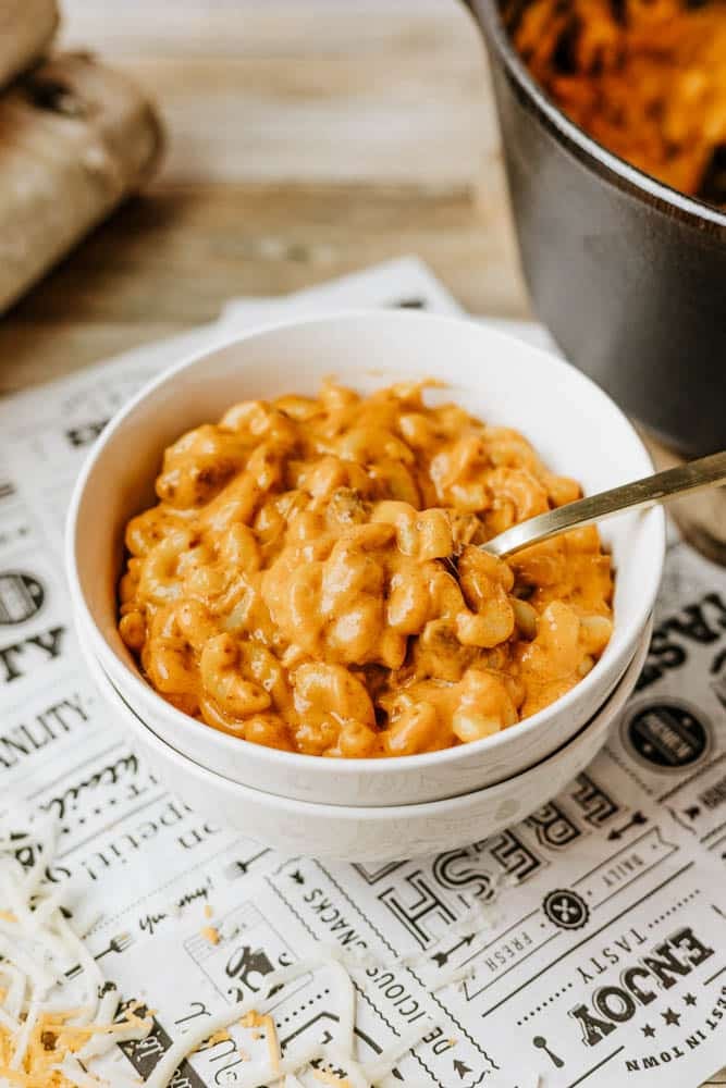 A white bowl filled with creamy macaroni and cheese with a metal spoon, placed on a sheet of printed paper with shredded cheese scattered around. A pot, perfect for making dutch oven chili mac, is partially visible in the background.
