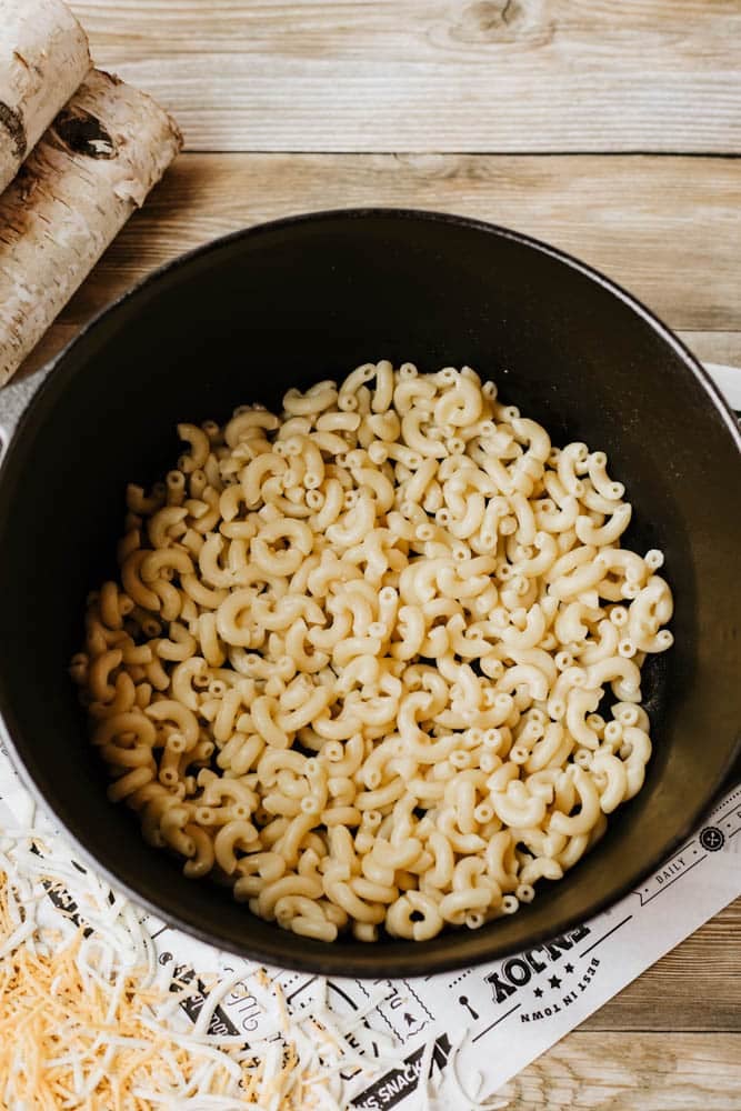 A black pot filled with cooked elbow macaroni sits on a wooden table, alongside shredded cheese and a decorative paper underneath—perfect for serving hearty Dutch oven chili mac.