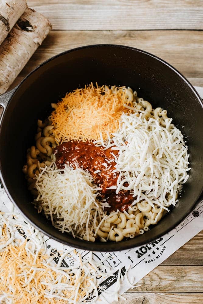 A black pot of dutch oven chili mac, filled with cooked elbow macaroni, topped with vibrant red marinara sauce and three piles of shredded cheese, sits on newspaper on a wooden table.
