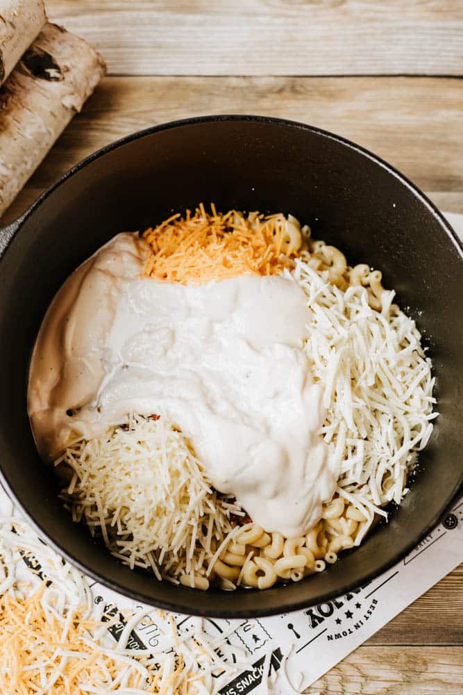 A black pot, similar to those used for dutch oven chili mac, is filled with uncooked pasta, shredded cheese, and a creamy white sauce, all placed on a wooden surface with paper underneath.
