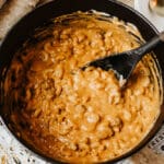 A black pot of creamy, cheesy macaroni and cheese is being stirred with a black spatula, resembling the comfort of dutch oven chili mac. Shredded cheese and a white plate with a spoon are nearby on a wooden surface.