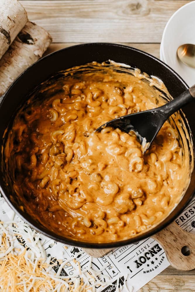 A black pot of creamy, cheesy macaroni and cheese is being stirred with a black spatula, resembling the comfort of dutch oven chili mac. Shredded cheese and a white plate with a spoon are nearby on a wooden surface.
