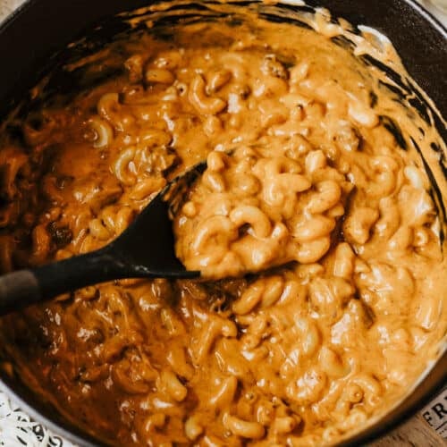 A black pot filled with creamy macaroni and cheese is being stirred with a black spoon. The cheesy sauce coats the elbow pasta, hinting at a delicious dutch oven chili mac, with some shredded cheese visible on the table nearby.