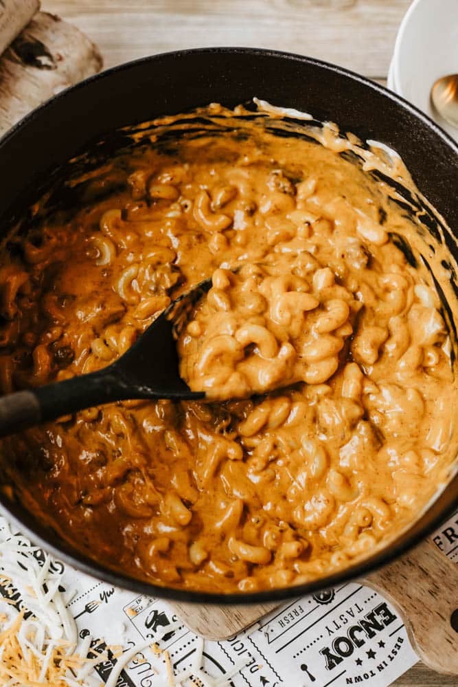 A black pot filled with creamy macaroni and cheese is being stirred with a black spoon. The cheesy sauce coats the elbow pasta, hinting at a delicious dutch oven chili mac, with some shredded cheese visible on the table nearby.
