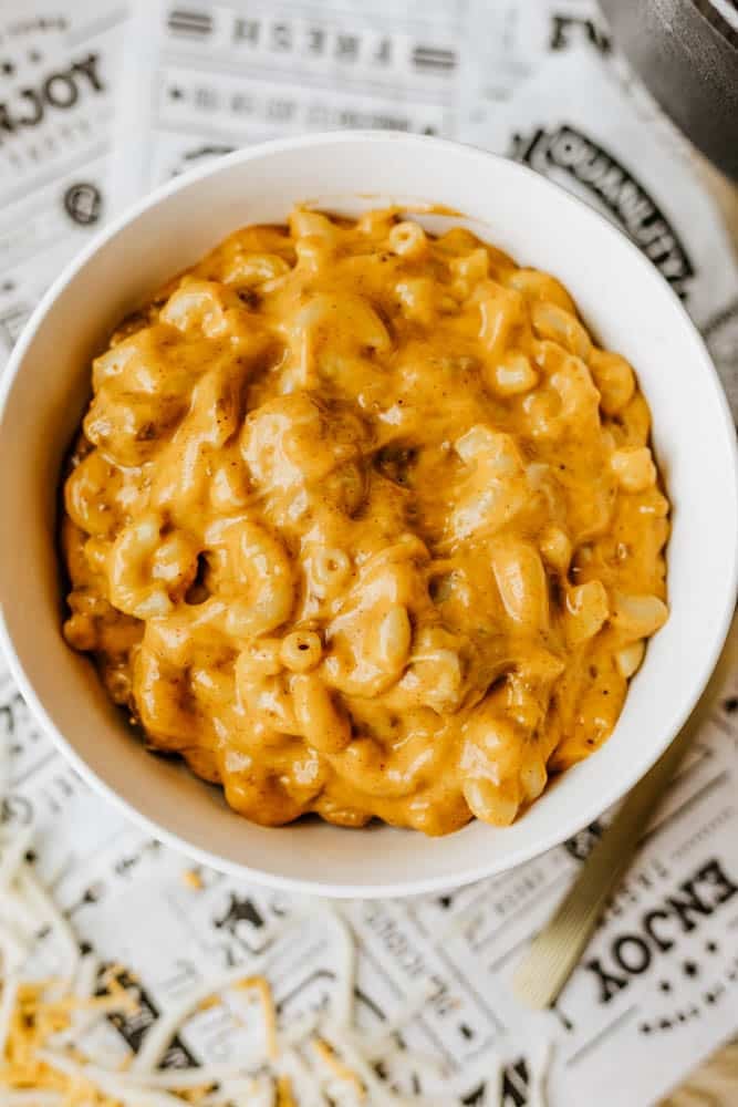 A white bowl filled with creamy, cheesy macaroni and cheese sits on a printed paper surface, with shredded cheese scattered nearby—a cozy dish reminiscent of hearty Dutch oven chili mac.