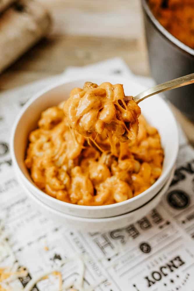 A close-up of creamy macaroni and cheese, inspired by dutch oven chili mac, with a fork lifting a cheesy, saucy bite. The food sits on a sheet of printed paper, and the dish looks rich and appetizing.