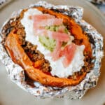 A foil packet sweet potato, split open and topped with savory ground beef, cottage cheese, guacamole, and pickled onions, served on a beige plate.