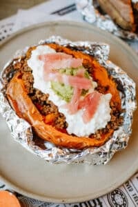 A foil packet sweet potato, split open and topped with savory ground beef, cottage cheese, guacamole, and pickled onions, served on a beige plate.