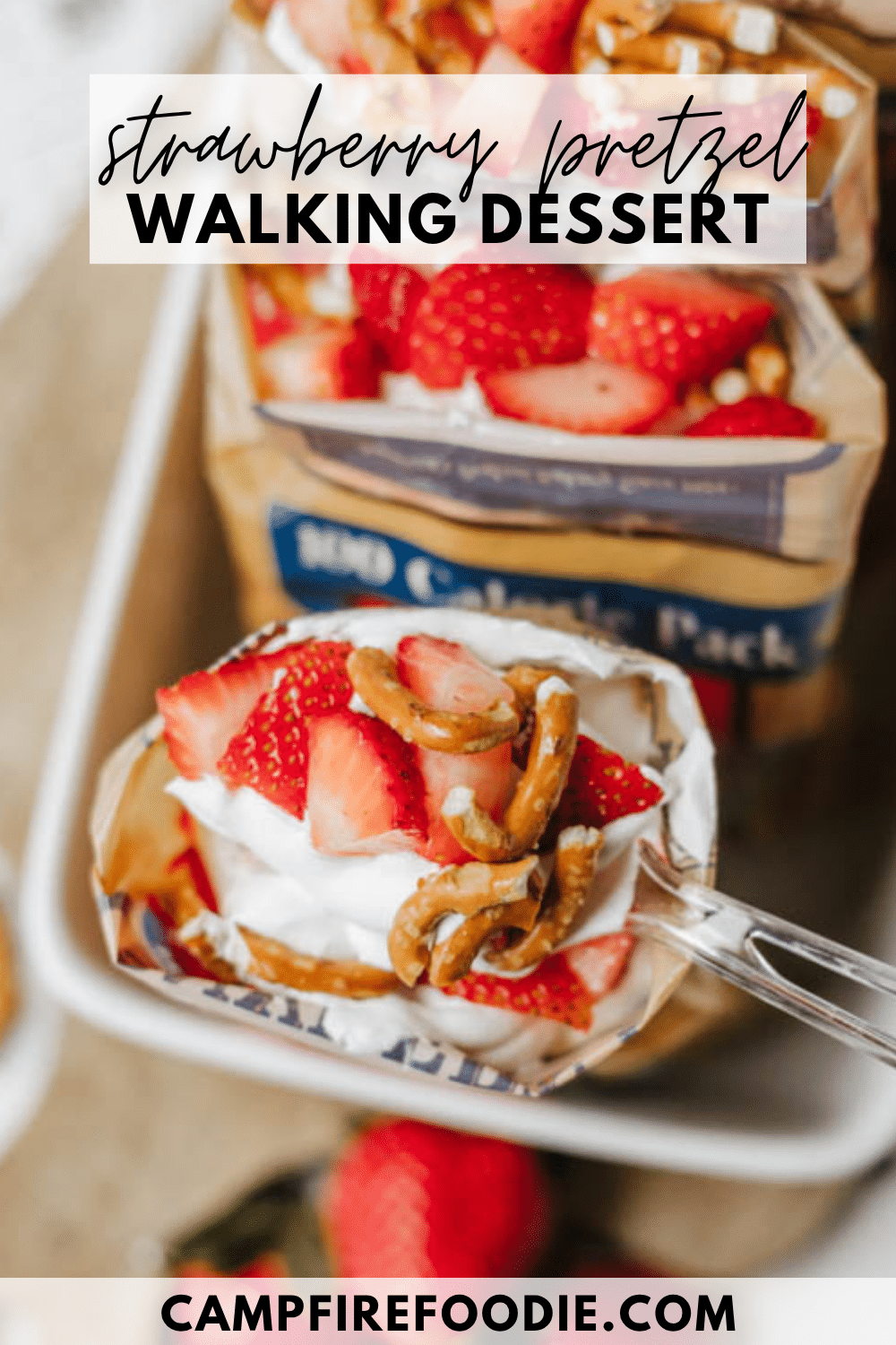 A spoonful of strawberry pretzel walking dessert with whipped cream, sliced strawberries, and pretzels, above an open snack bag. Text reads strawberry pretzel walking dessert and campfirefoodie.com.