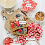 Various dessert ingredients are arranged on a white surface: bowls of pudding, pretzels, sliced strawberries, and Cool Whip, along with Jello cups and two bags of pretzel sticks on a wooden board with a red checkered cloth.