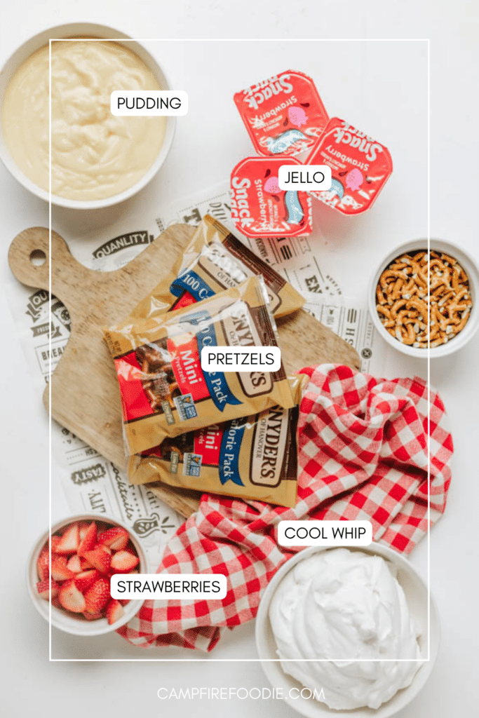 Various dessert ingredients are arranged on a white surface: bowls of pudding, pretzels, sliced strawberries, and Cool Whip, along with Jello cups and two bags of pretzel sticks on a wooden board with a red checkered cloth.