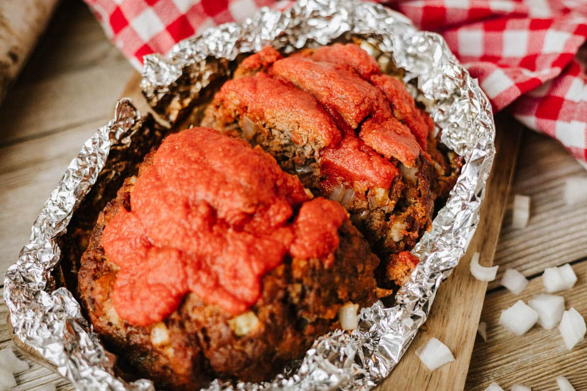 A foil packet meatloaf recipe topped with tomato sauce sits on a wooden board, wrapped in aluminum foil, with chopped onions scattered nearby and a red-and-white checkered cloth in the background.