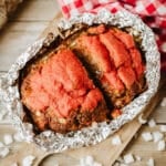 A foil packet meatloaf recipe sits in tomato sauce on a wooden cutting board, surrounded by diced onions and a red and white checkered cloth.
