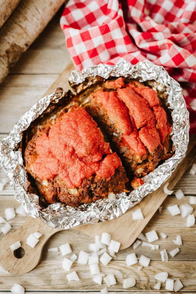 A foil packet meatloaf recipe sits in tomato sauce on a wooden cutting board, surrounded by diced onions and a red and white checkered cloth.
