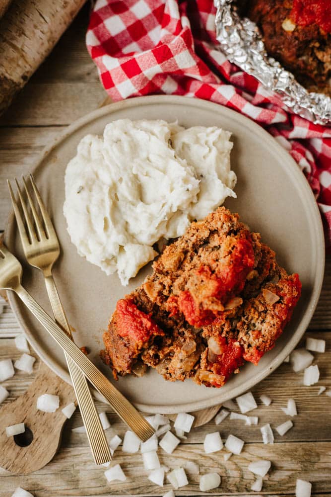 A plate with two slices of foil packet meatloaf recipe topped with tomato sauce sits beside mashed potatoes and two gold forks. Diced onions are scattered on the wooden table, near a red and white checkered cloth.