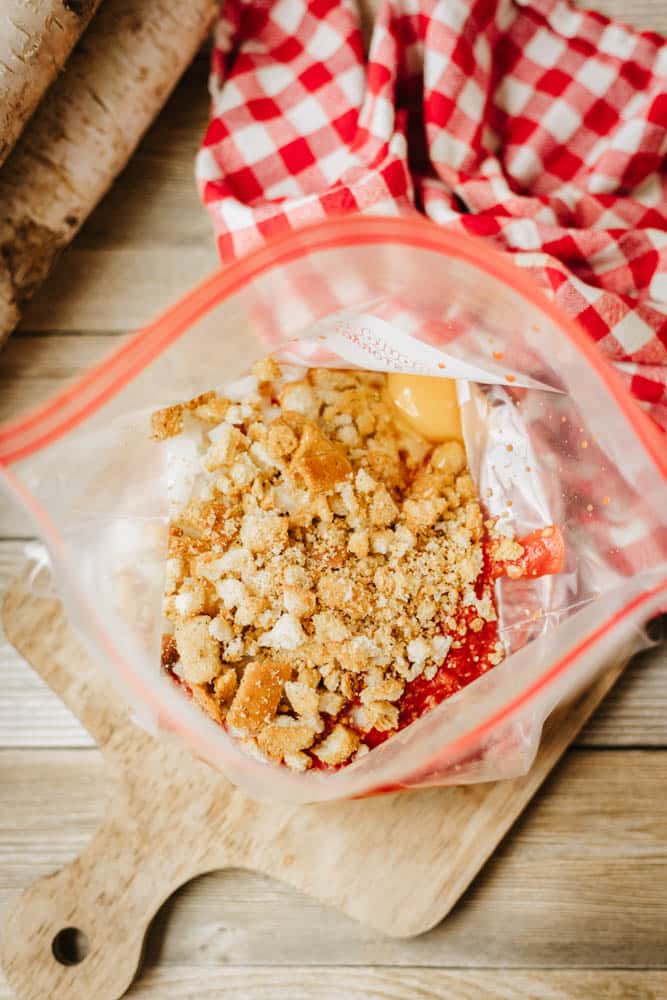 A clear plastic bag filled with crumbled crackers, shredded cheese, and an orange sauce sits on a wooden surface near a red and white checkered cloth—perfect prep ingredients for a tasty foil packet meatloaf recipe.