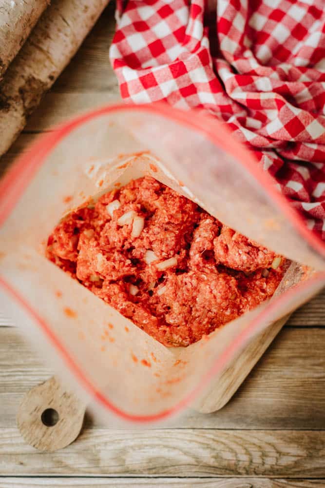 A plastic bag filled with seasoned ground meat and chopped onions, perfect for a foil packet meatloaf recipe, sits on a wooden surface next to a red and white checkered cloth.
