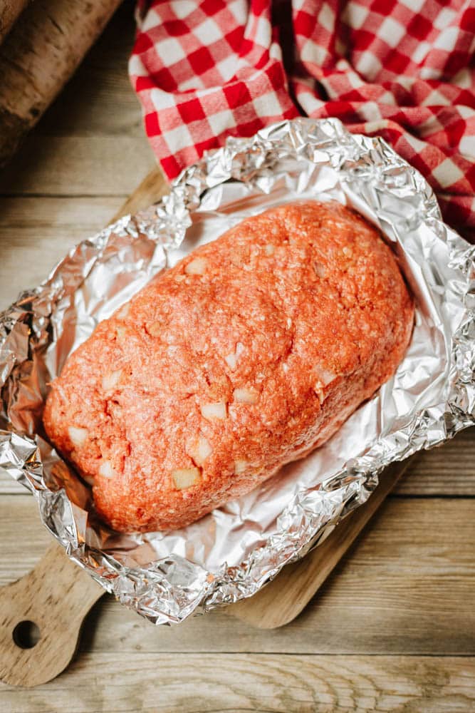 A raw foil packet meatloaf recipe mixture with visible diced onions sits on a foil-lined baking dish, placed on a wooden surface next to a red and white checkered cloth.